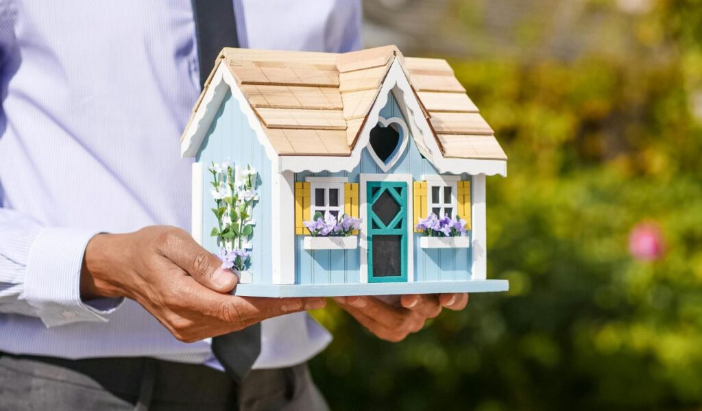 "Person in business attire holding a decorative model house, symbolizing renting out your home while traveling."