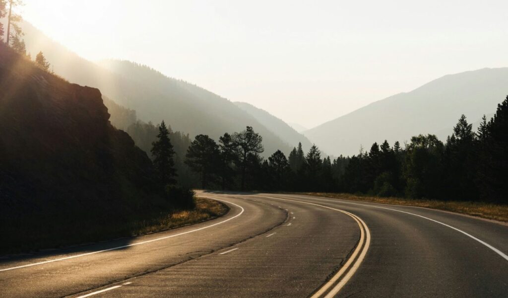 A winding, empty road curves through a mountainous forest landscape during sunrise or sunset, with soft light casting long shadows across the pavement.