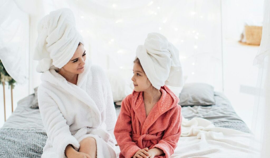 Mother and daughter in bathrobes with towels wrapped on their heads, sitting on a bed and smiling at each other.