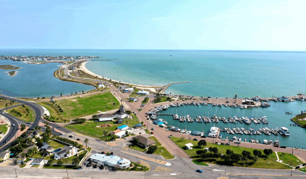 Aerial view of Rockport, Texas showing marina with docked boats, curving coastline, and beach meeting the Gulf of Mexico.