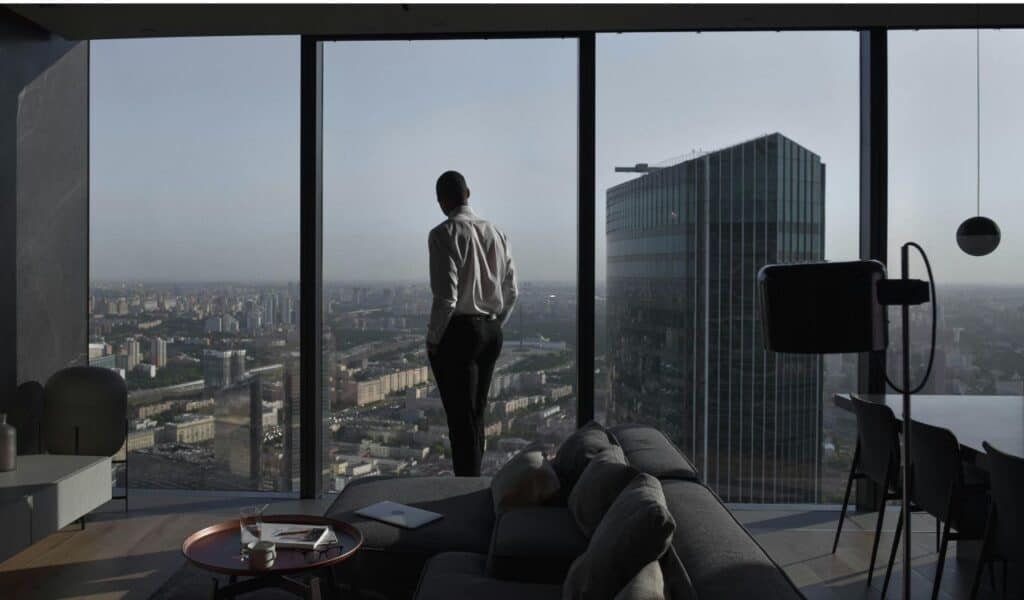 A man in a formal shirt stands by the floor-to-ceiling windows of a modern high-rise suite, overlooking a sprawling cityscape with skyscrapers and urban buildings at dusk.