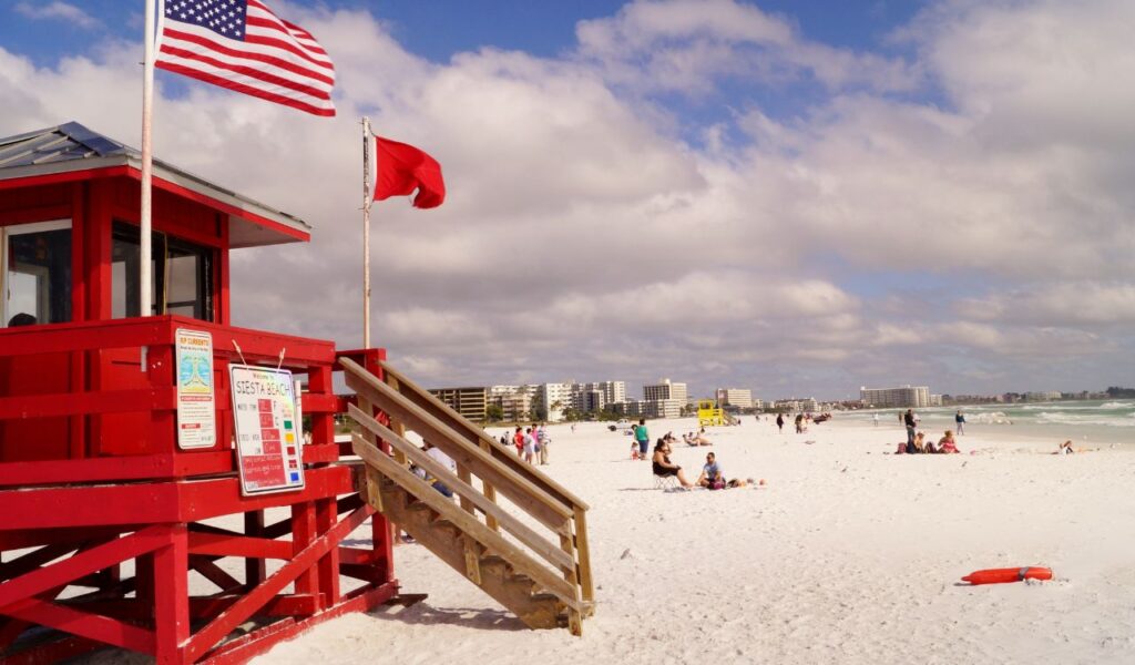 Red lifeguard tower with warning flags and beach safety signs on a busy Florida beach with swimmers and buildings in the distance.
