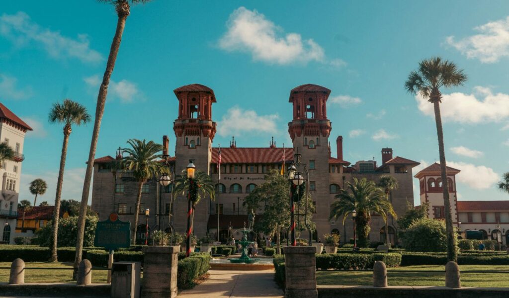 Historic building in St. Augustine, Florida, with twin towers, palm trees, manicured gardens, and a central fountain under a clear blue sky.