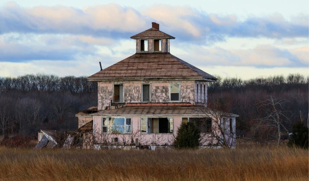 An abandoned, weathered house with peeling paint and broken windows sits in a field of tall grass, backed by leafless trees and a cloudy sky.