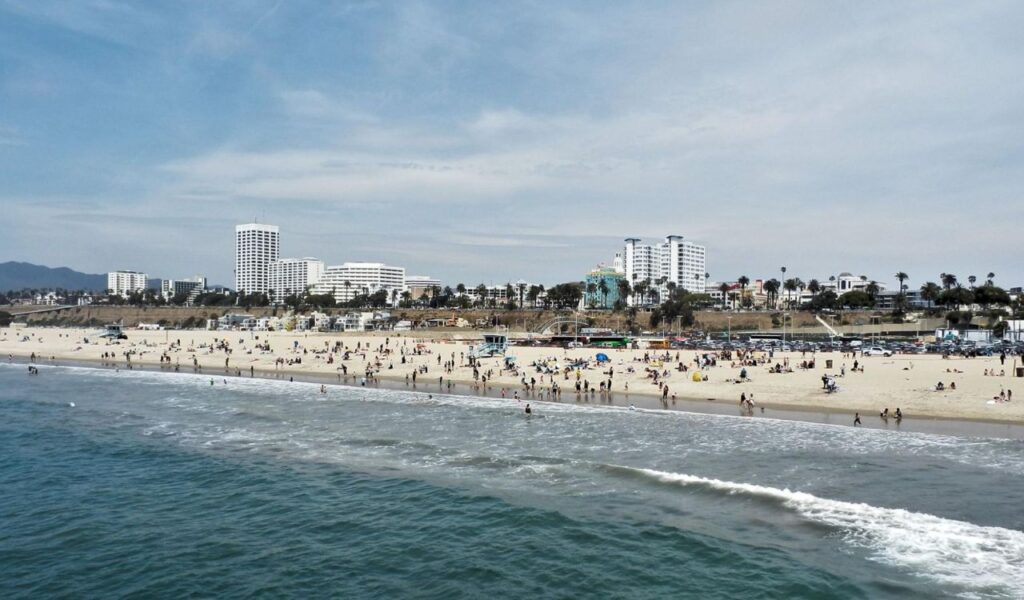 View of Santa Monica Beach with people on the sand, ocean waves, and city buildings in the background.