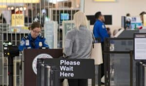 A TSA officer assists a passenger at an airport security checkpoint with a 'Please Wait Here' sign in view."