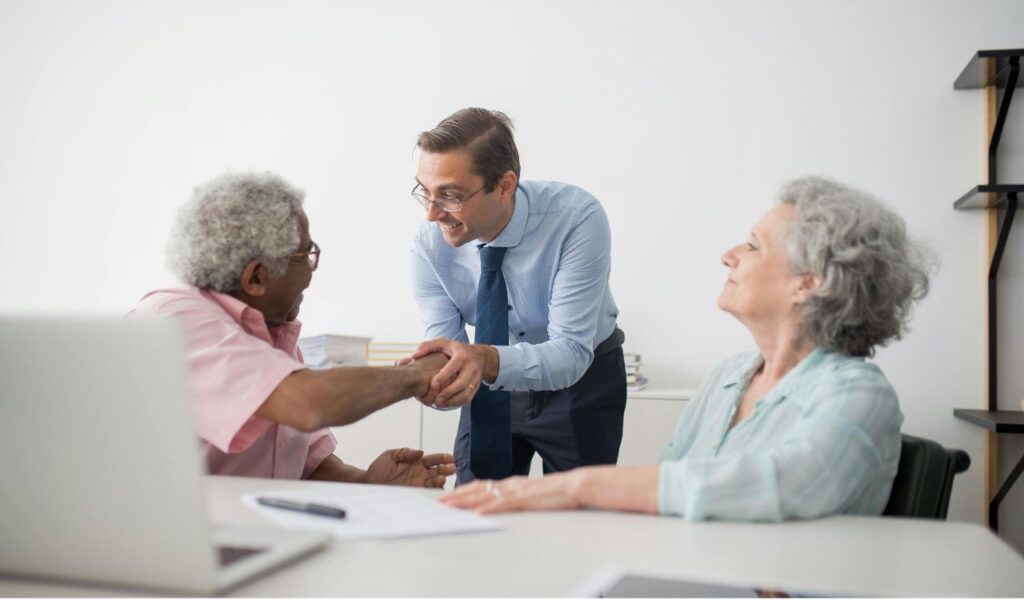 An elderly couple meets with an insurance professional to discuss travel insurance options, shaking hands and making a decision.