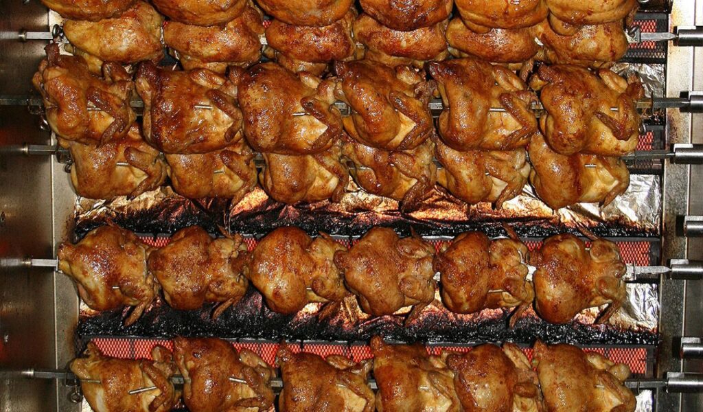 Rows of whole rotisserie chickens cooking on a commercial spit, with golden-brown, crispy skin glistening under the heat.