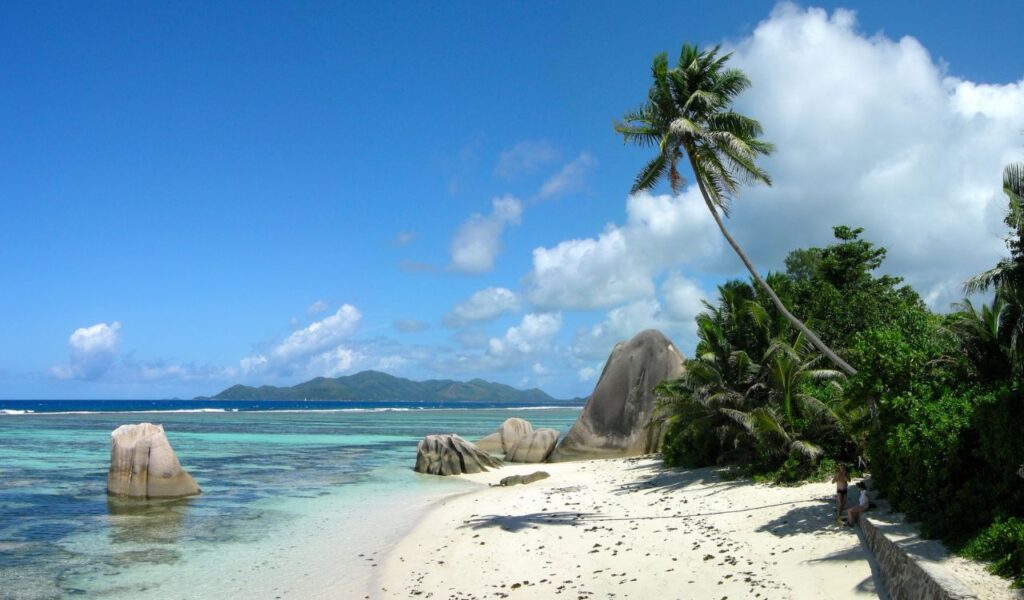 Tropical beach in Seychelles with giant granite boulders, turquoise water, and leaning palm trees under sunny skies.