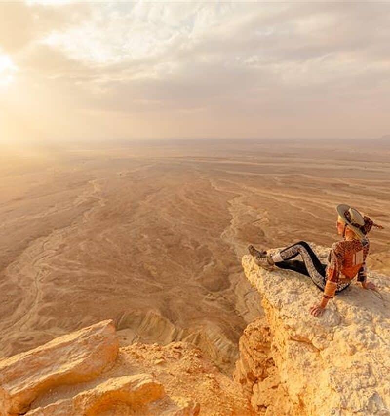 Female tourist wearing hat and sunglasses sitting on rock admiring sunset view of Tuwaiq escarpment, edge of the world, in Riyadh, Saudi Arabia, a breathtaking desert landscape