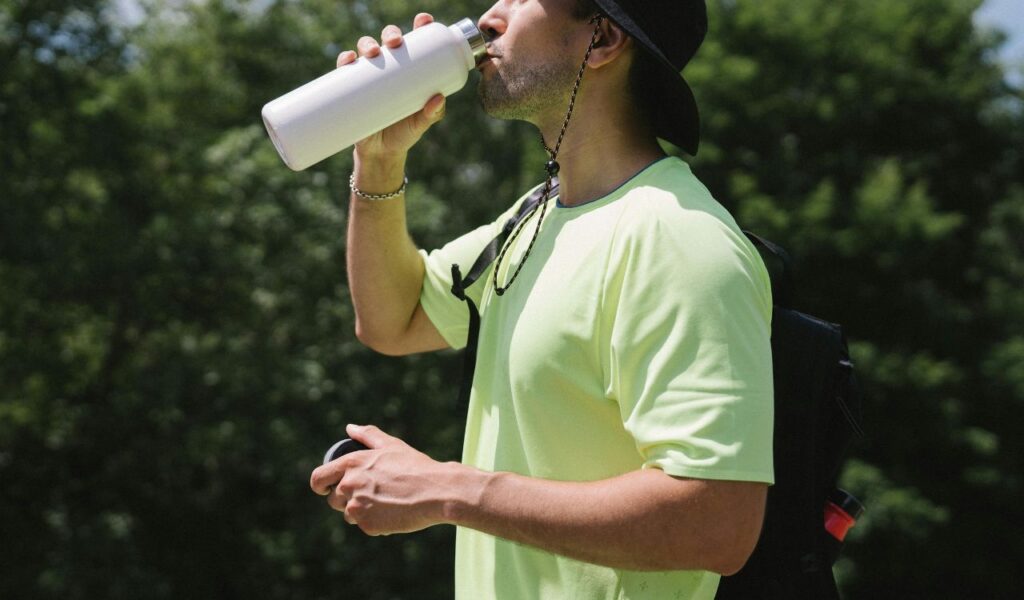 Man wearing a neon shirt and black hat drinking from a reusable water bottle outdoors, staying hydrated in warm weather.
