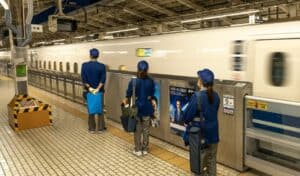 Workers in blue uniforms standing in an orderly line on a Japanese train platform as a high-speed Shinkansen train passes by, showcasing Japan’s culture of organized queuing.