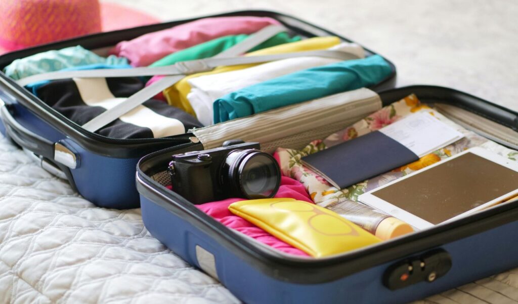 An open suitcase neatly packed with clothes, a camera, sunglasses case, tablet, passport, and travel documents, placed on a bed, ready for a trip.