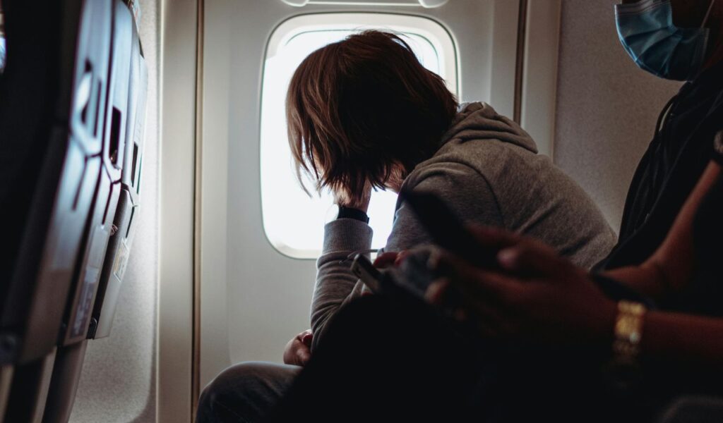 Passenger in hoodie resting against airplane window, appearing tired while seated during long-haul flight.
