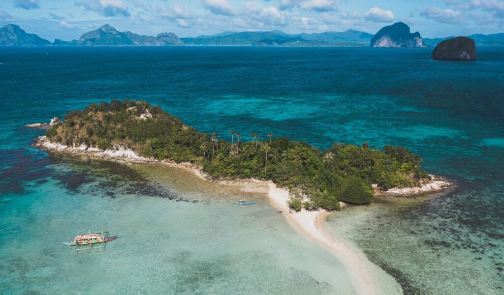 A remote tropical island surrounded by turquoise waters and coral reefs, with boats nearby and dense greenery covering the land, suggesting restricted or untouched access.