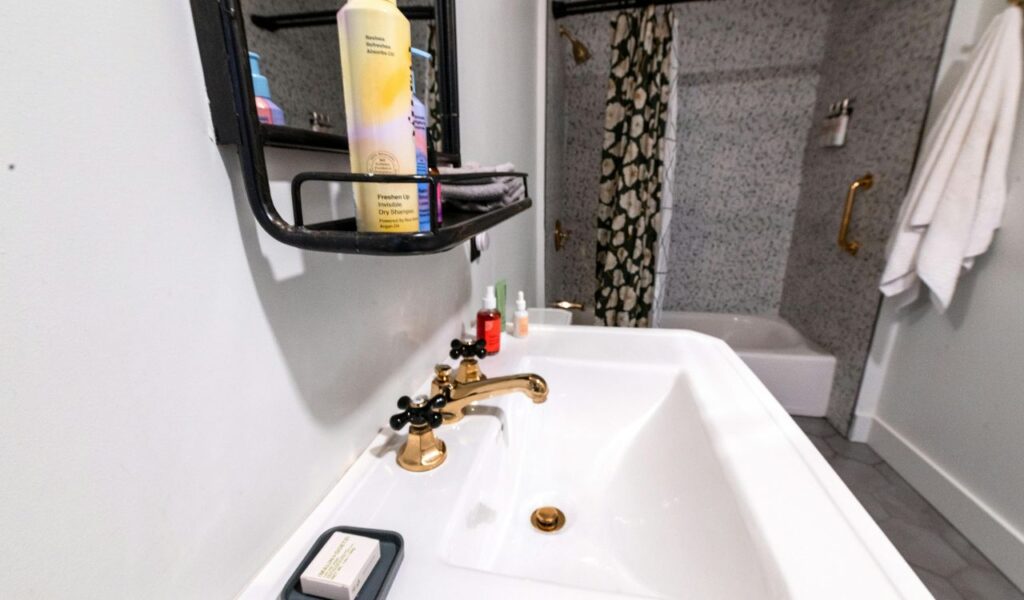 Bathroom sink with gold faucet, soap dish, toiletries, and a bathtub with a floral shower curtain in the background.