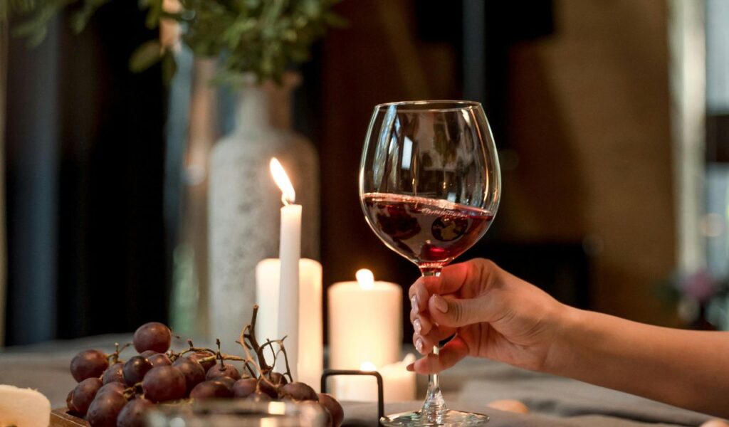 A hand holding a glass of red wine beside lit candles and a bunch of grapes, creating a cozy, intimate setting.
