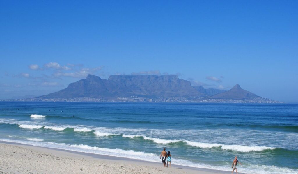 Couple walking along Cape Town beach with Table Mountain in the background under clear blue sky – scenic South Africa coastline.