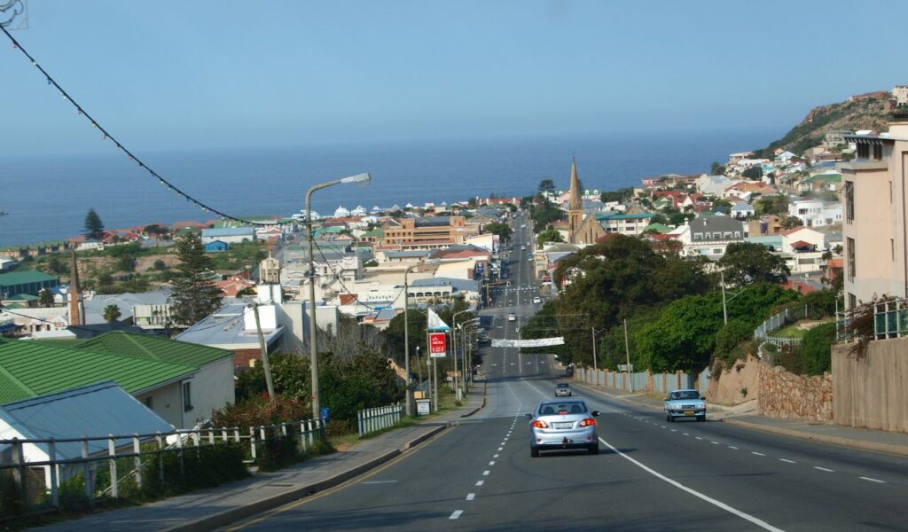 Coastal town view from the Garden Route in South Africa, with ocean in the background and vehicles driving downhill.