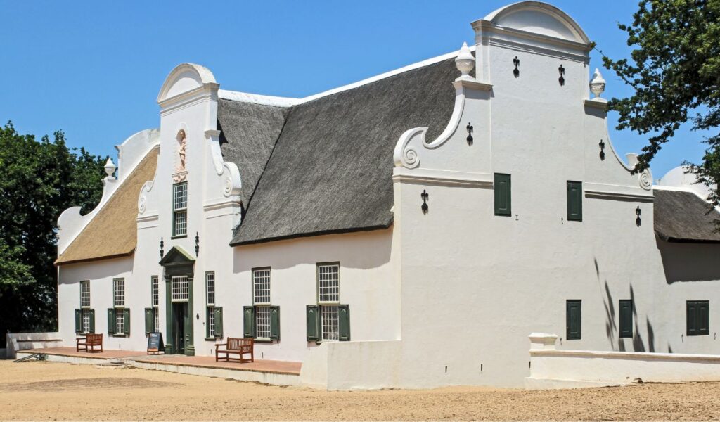 A white Cape Dutch-style building with ornate gables, green shutters, and a thatched roof, set under a clear blue sky with benches out front.