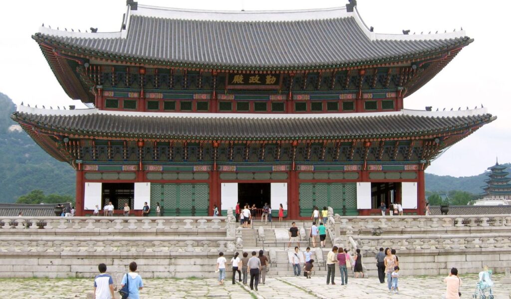 Traditional Korean palace building with a multi-tiered, curved roof and colorful wooden detailing, as visitors explore its stone steps and courtyard.