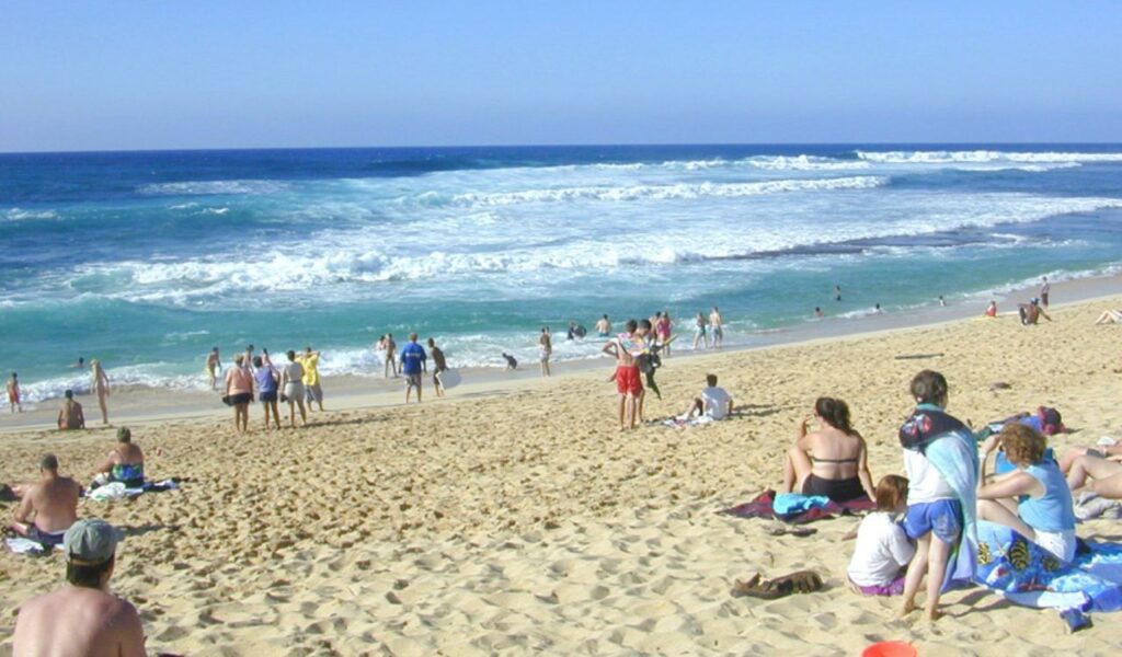 People relaxing and playing at a sandy beach in Spain with waves crashing in the background under a clear blue sky.