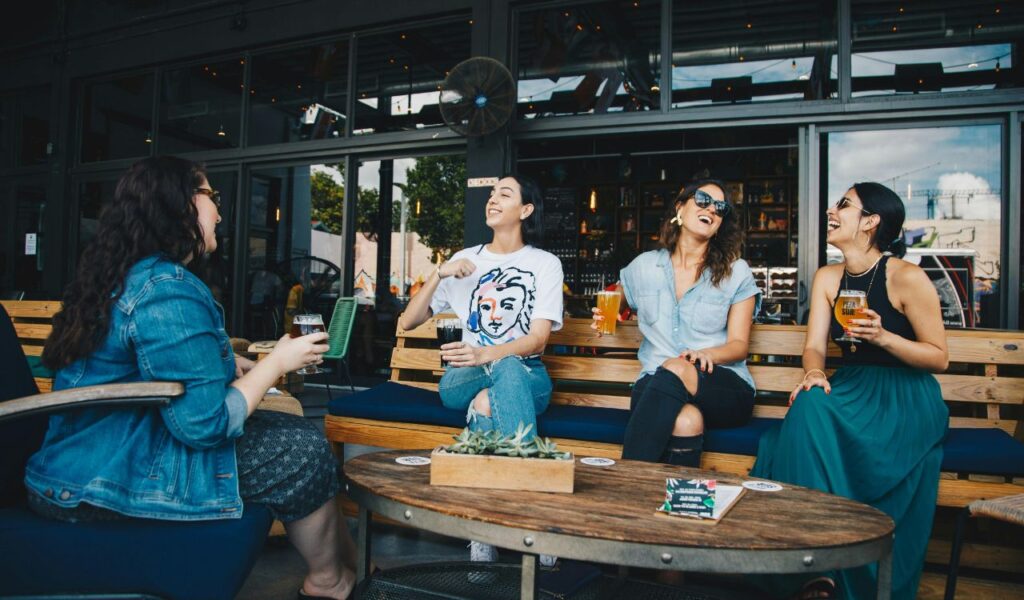 Four women sitting outside a bar or café, laughing and holding drinks, suggesting a social and relaxed atmosphere that could be relevant to themes like nightlife, dating, or potential drink-related scams.