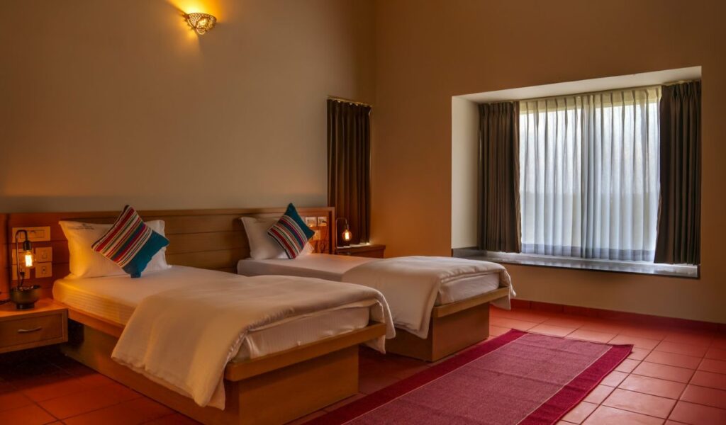 A cozy hotel room with two neatly made single beds, colorful pillows, warm lighting, terracotta tile flooring, and a large window with sheer curtains.