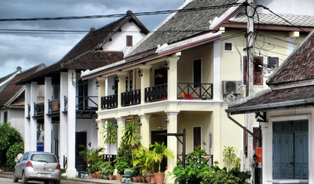 Colorful colonial-style guest houses with balconies and potted plants lining a quiet street in Luang Prabang, Laos, under a cloudy sky.