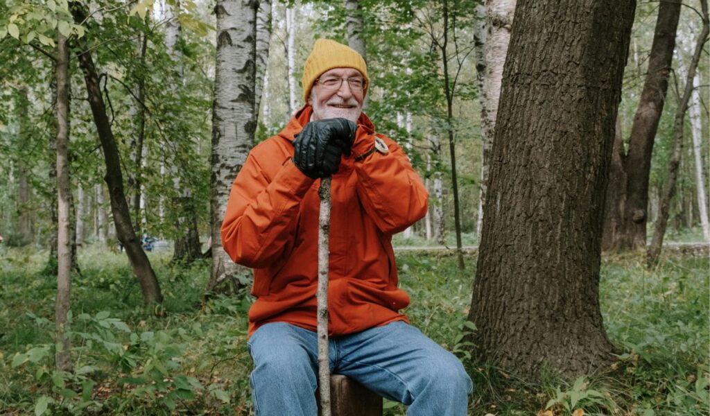 An elderly man sitting on a tree stump in the forest, smiling while wearing an orange jacket, yellow beanie, and black gloves, holding a walking stick.