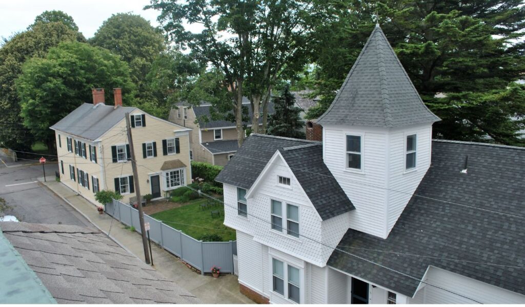 Charming street view of Stonington, Maine with historic wooden homes, a pointed turret, and lush green trees lining the road.