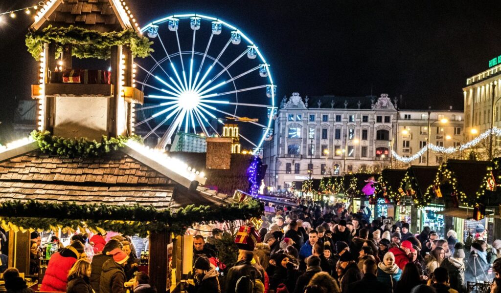 Crowds of people enjoying the festive Strasbourg Christmas Market at night, with wooden stalls decorated with lights and greenery, and a brightly lit Ferris wheel in the background.