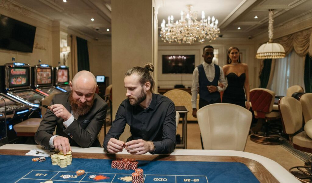 Two men sitting at a casino gambling table with poker chips, while a well-dressed couple approaches in the background, suggesting a high-stakes gaming environment.