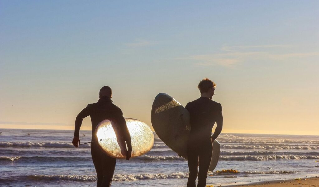 Two surfers walking along the beach at sunset, carrying their surfboards and heading towards the ocean.