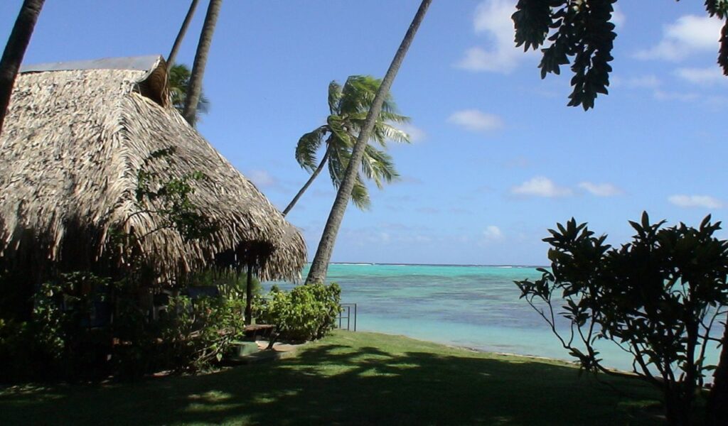 Traditional thatched hut on a Tahiti beach with palm trees, turquoise lagoon, and clear blue sky in the background.