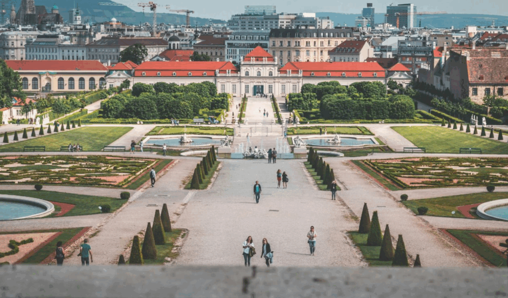 Visitors stroll through a grand European palace garden with symmetrical fountains, trimmed hedges, and a city skyline rising in the background.