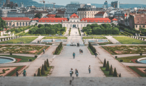 Visitors stroll through a grand European palace garden with symmetrical fountains, trimmed hedges, and a city skyline rising in the background.