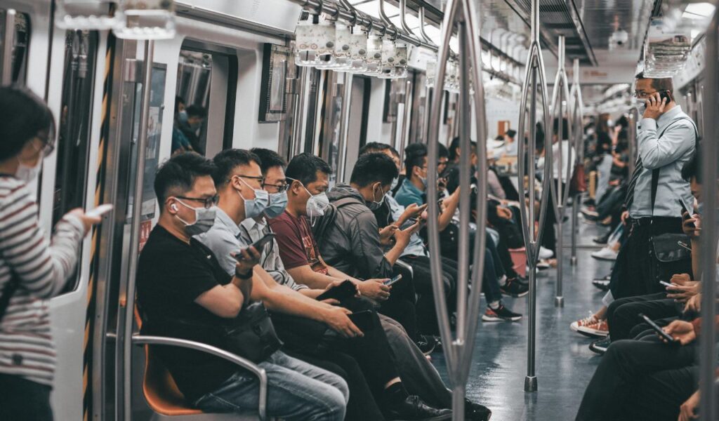 Passengers sit quietly in a subway train, most wearing masks and using their phones, while one man stands talking on his phone, contrasting with the calm and silent atmosphere expected in public transport.