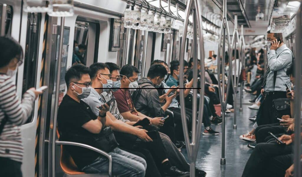 Passengers seated quietly in a subway train, most wearing masks and using their phones, with one standing man talking on his phone, highlighting the contrast to the calm and silent environment typical of public transport.