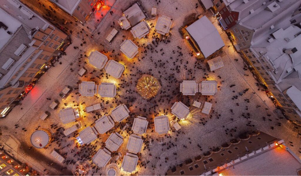 Aerial view of Tallinn Christmas Market in Estonia, with snow-covered stalls arranged in a circular pattern around a glowing central Christmas tree, surrounded by historic buildings.