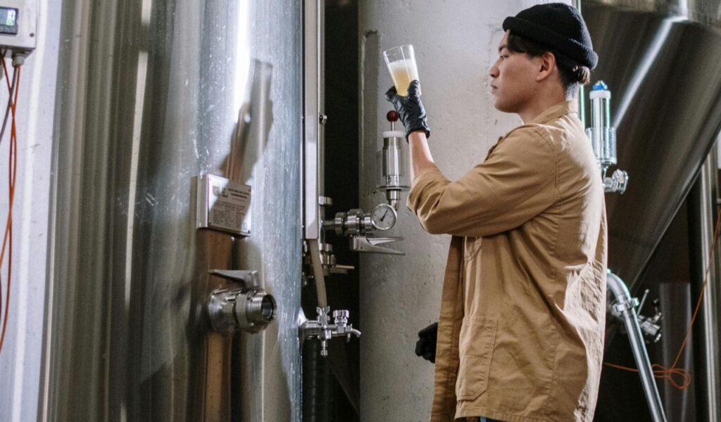 A brewer in a beige jacket and black beanie examines a glass of freshly poured beer in a brewery, standing beside large stainless steel brewing tanks.