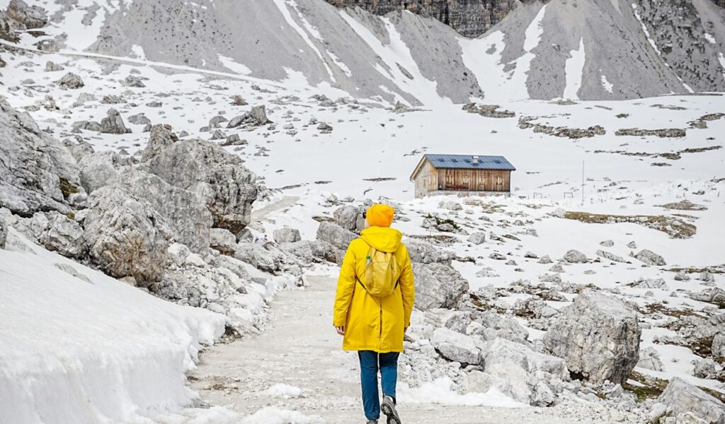 Solo hiker in yellow jacket walking through snowy mountain trail toward rocky peaks.