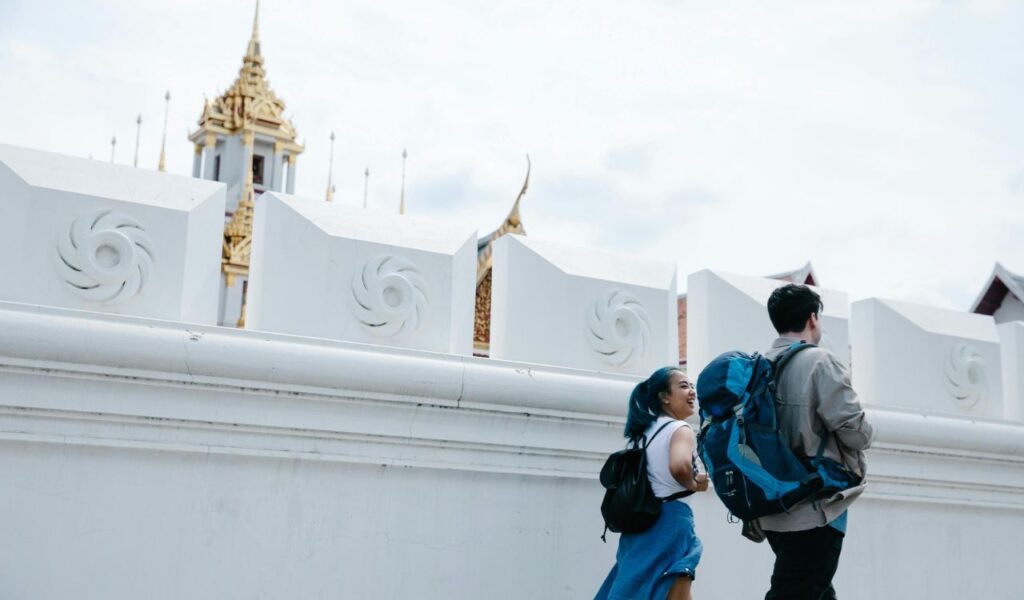 Two backpackers walk beside a white temple wall in Thailand, with golden rooftops and spires visible above the structure.