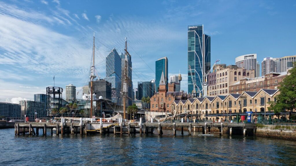 Sydney waterfront with historic buildings, modern skyscrapers, and boats docked along the pier under a clear blue sky.