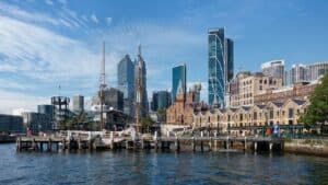 Sydney waterfront with historic buildings, modern skyscrapers, and boats docked along the pier under a clear blue sky.