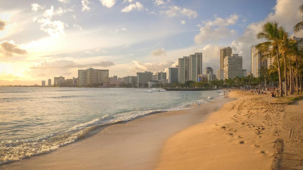 A serene view of Waikiki Beach at sunset, with golden sand, gentle waves, palm trees, and Honolulu’s skyline in the background.