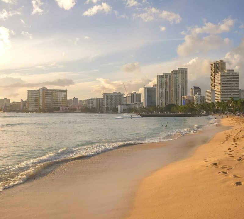 A serene view of Waikiki Beach at sunset, with golden sand, gentle waves, palm trees, and Honolulu’s skyline in the background.