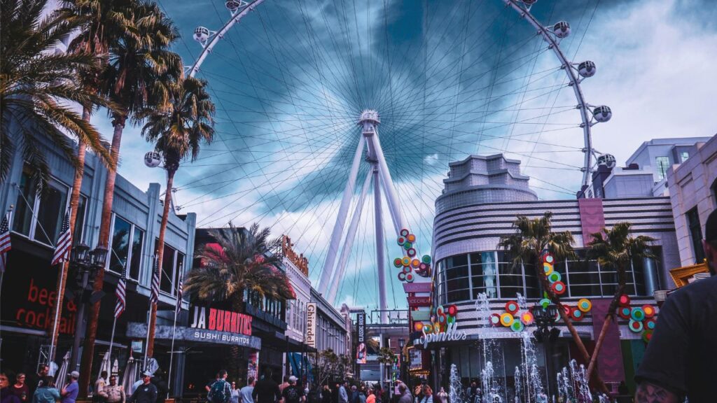 Crowded street view at an amusement area with palm trees, colorful storefronts, fountains, and a giant Ferris wheel towering in the background under a cloudy sky.