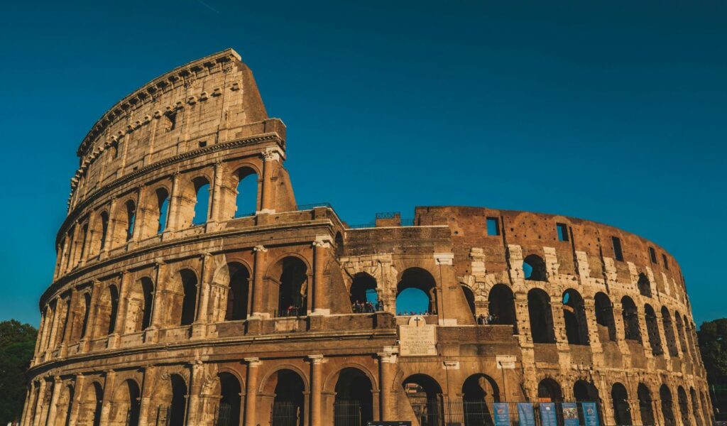 Ancient Roman Colosseum in Rome, Italy, under a clear blue sky with warm sunlight highlighting the stone arches.