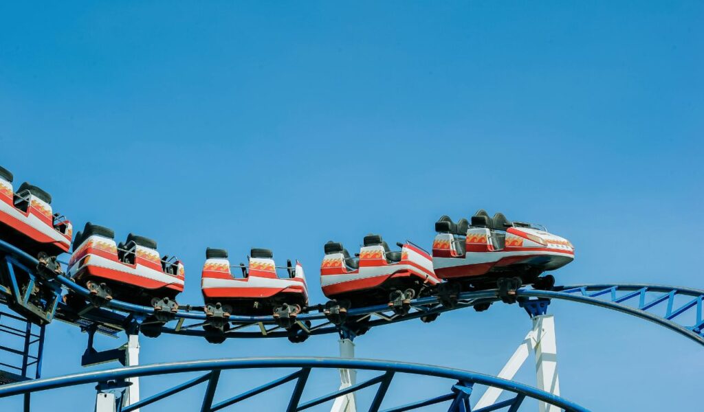 A red and white roller coaster train rides along a twisting blue steel track under a bright blue sky, with all seats empty and no passengers aboard.